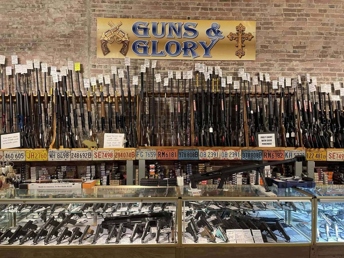 The main gun wall at Guns and Glory — rifles and shotguns lined up under the Guns & Glory sign on a brick wall, with handguns in the glass case below and ammunition stocked beneath.