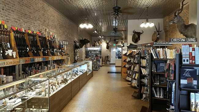 Looking down the length of Guns and Glory — guns on the left wall, Christian Living book section on the right, taxidermy mounts overhead.