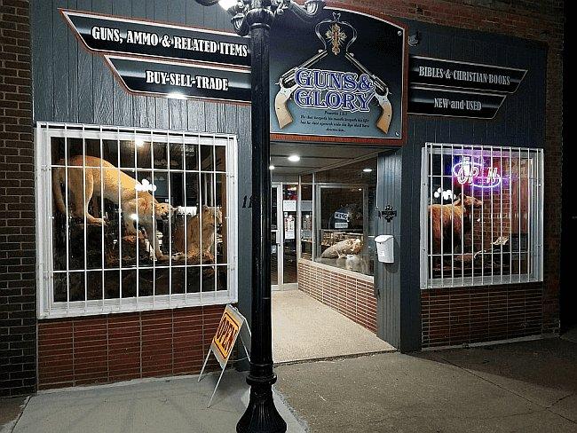 Guns and Glory storefront at night, with neon glow and taxidermy visible through the barred windows.